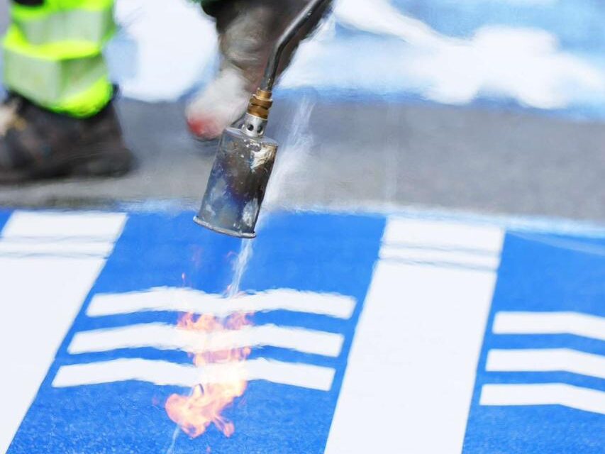 Close-up of a blue and white shield design made from Prismo preformed thermoplastic being applied to a street in Iceland using a gas torch.