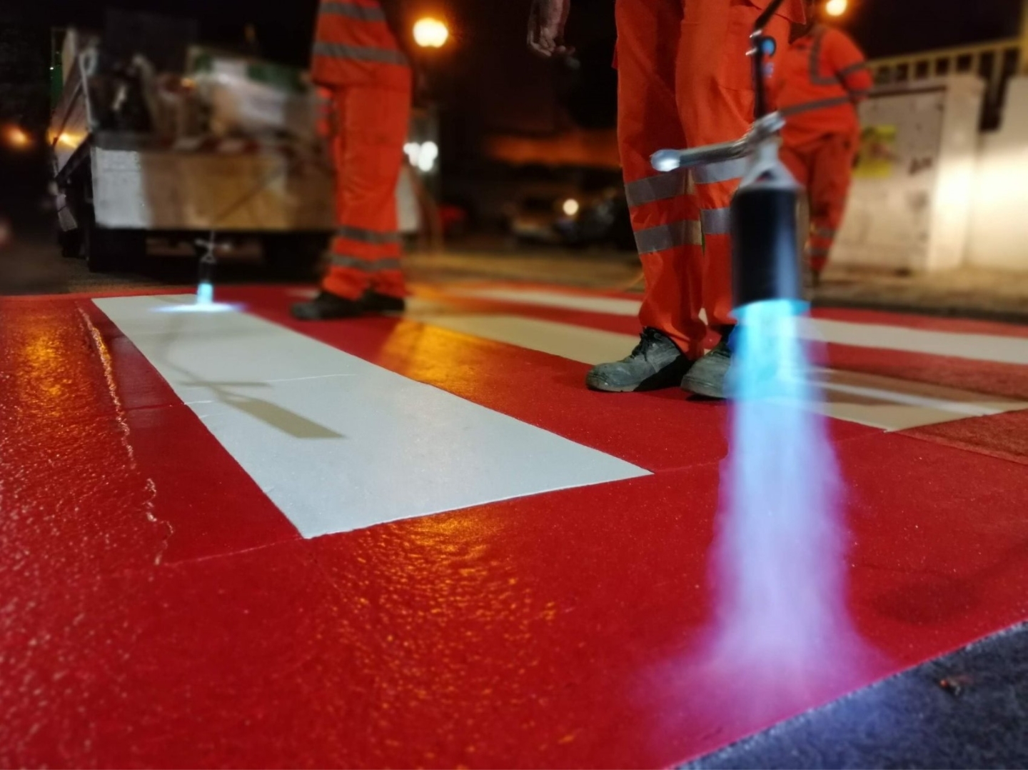 A close up of Prismo® Preformed Thermoplastic road marking being applied using a gas torch at a pedestrian crossing, including white crossing lines with a red background.
