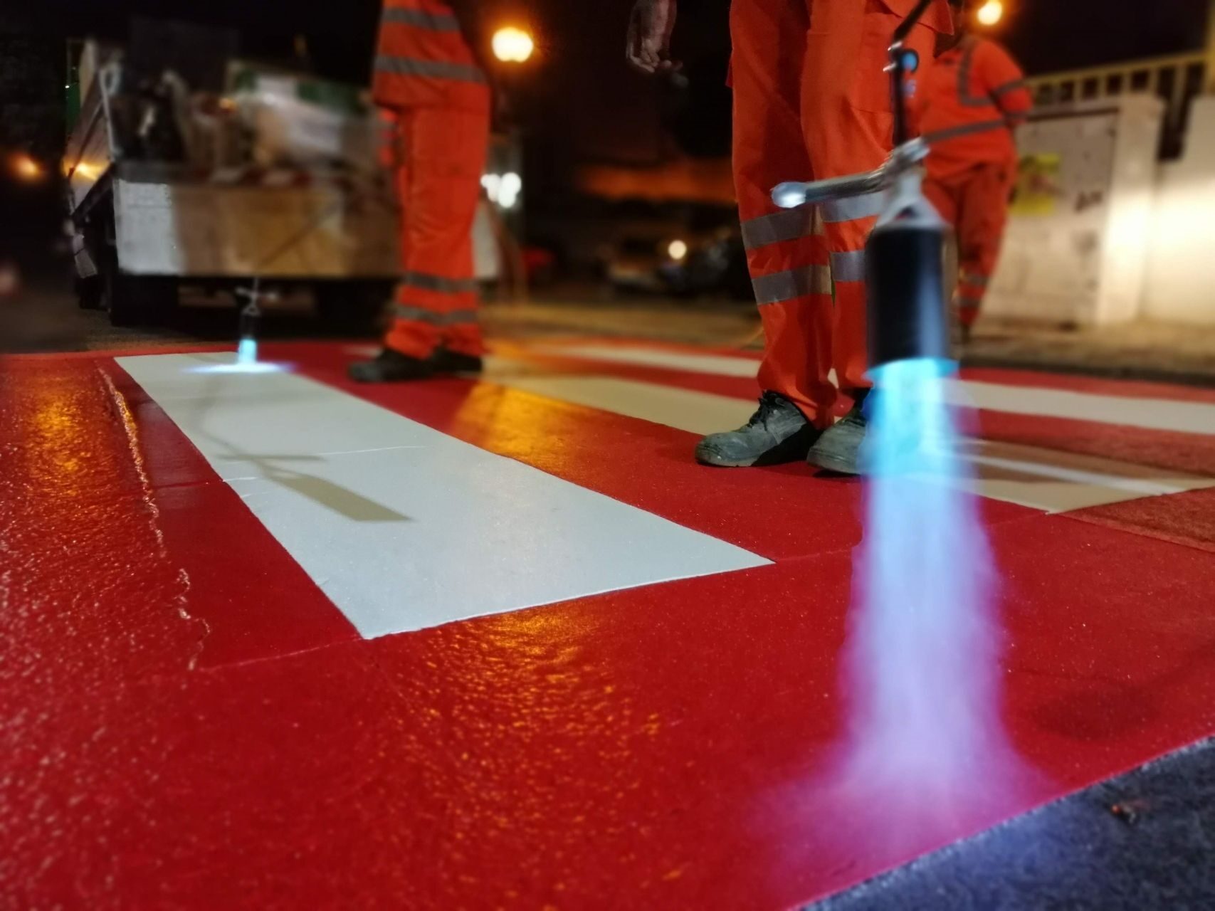 A close up of Prismo® Preformed Thermoplastic road marking being applied using a gas torch at a pedestrian crossing, including white crossing lines with a red background.