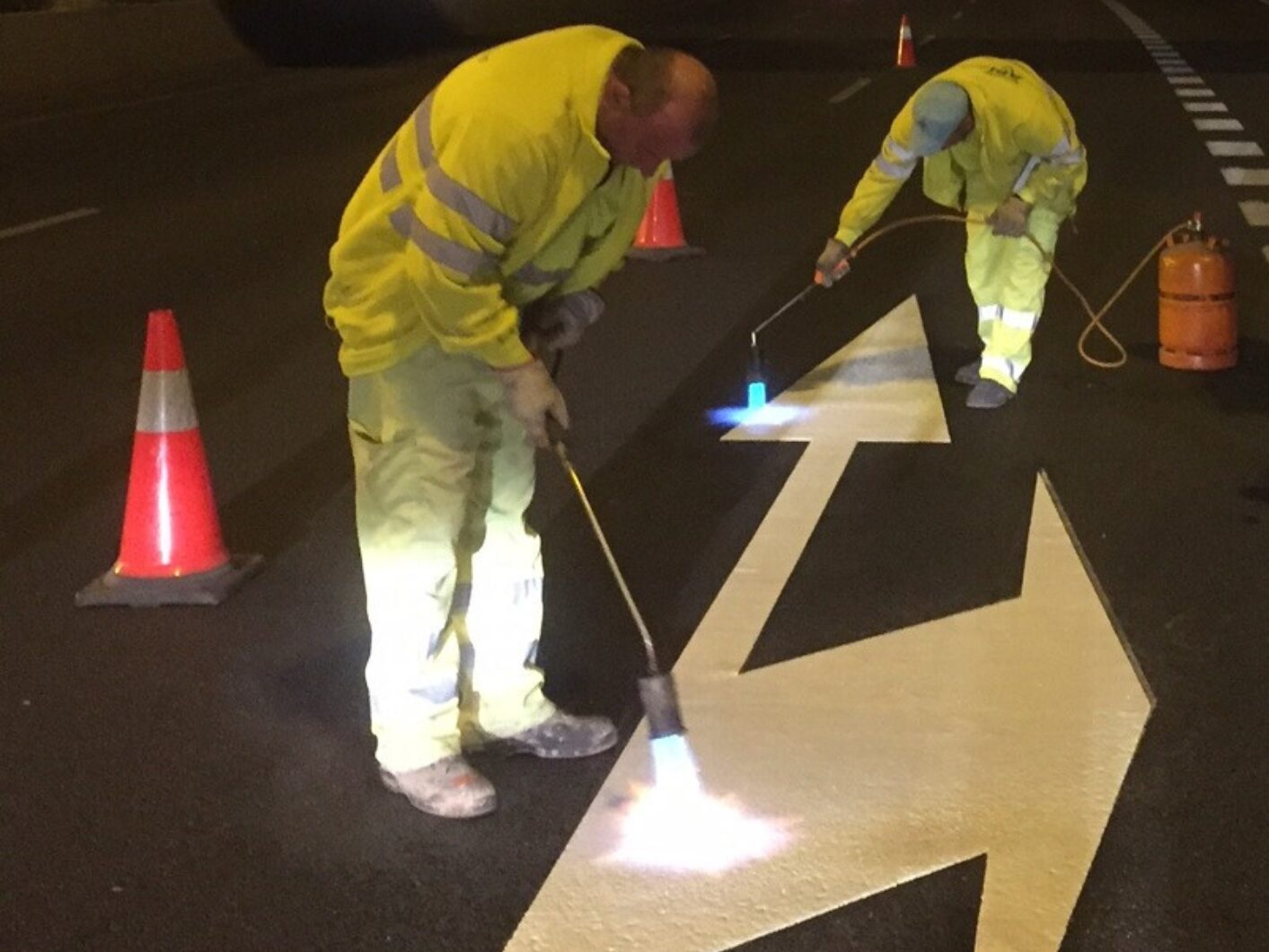 Prismo® Preformed Thermoplastic white directional arrow being applied by two contractors on a motorway using a gas torch at night.