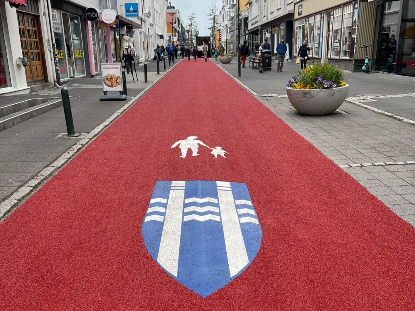 White parent and child symbol alongside a custom blue and white shield design on a red pedestrian street in Iceland, made from Prismo preformed thermoplastic road markings.