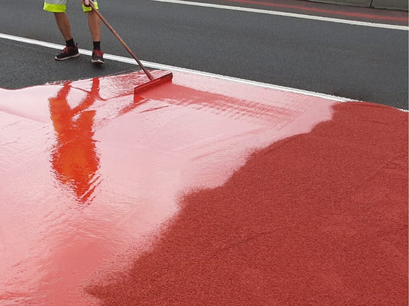 Contractor applying red Prismo Tyregrip high friction surfacing with a squeegee on a main road.