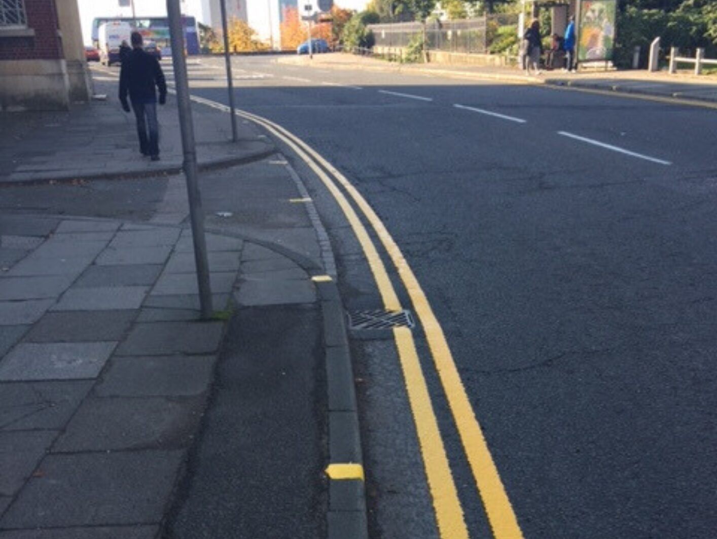 Prismo Guidemaster hot applied thermoplastic road markings are used in Salford for double yellow lines on the left side of the road surface, next to the pavement. A pedestrian walks on the pavement in the distance.