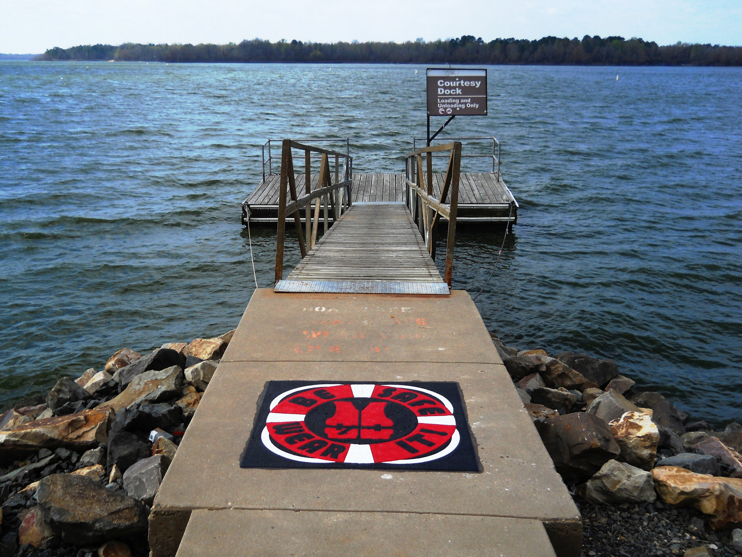 Decorative “Be Safe Wear It” life jacket safety marking in colours red, white and black made from Prismo preformed thermoplastic at the entrance of a lakeside dock.