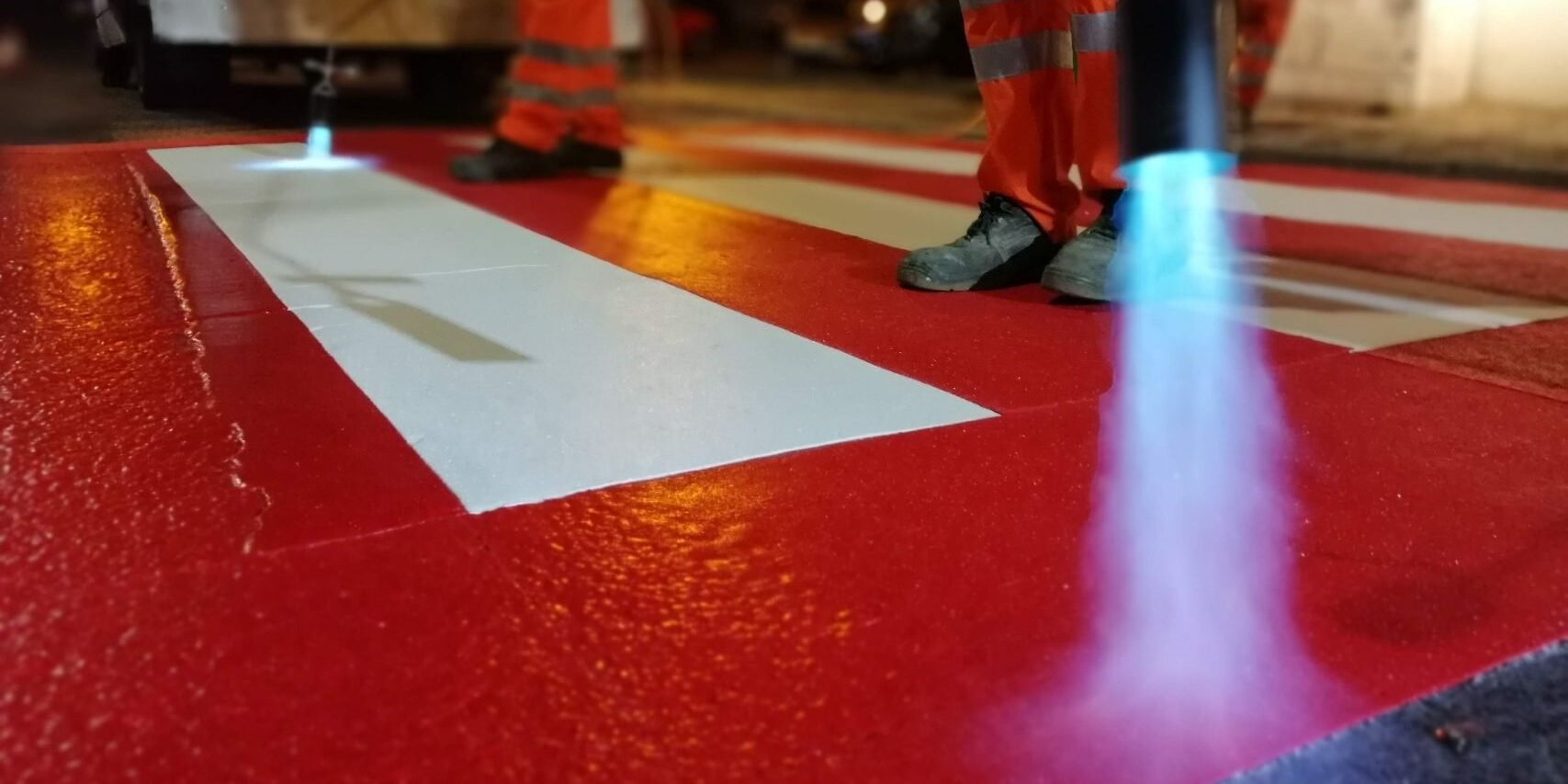 A close up of Prismo® Preformed Thermoplastic road marking being applied using a gas torch at a pedestrian crossing, including white crossing lines with a red background.