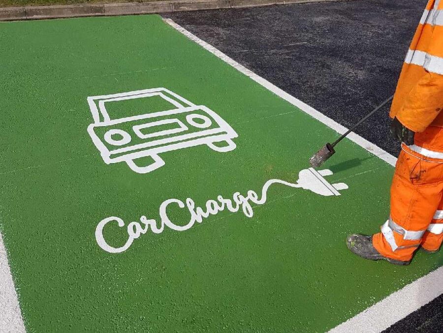 Prismo Preformed Thermoplastic white EV charging bay symbol being applied with a gas torch by a contractor in daylight. The EV bay symbol is a custom design on a refreshed green parking bay.