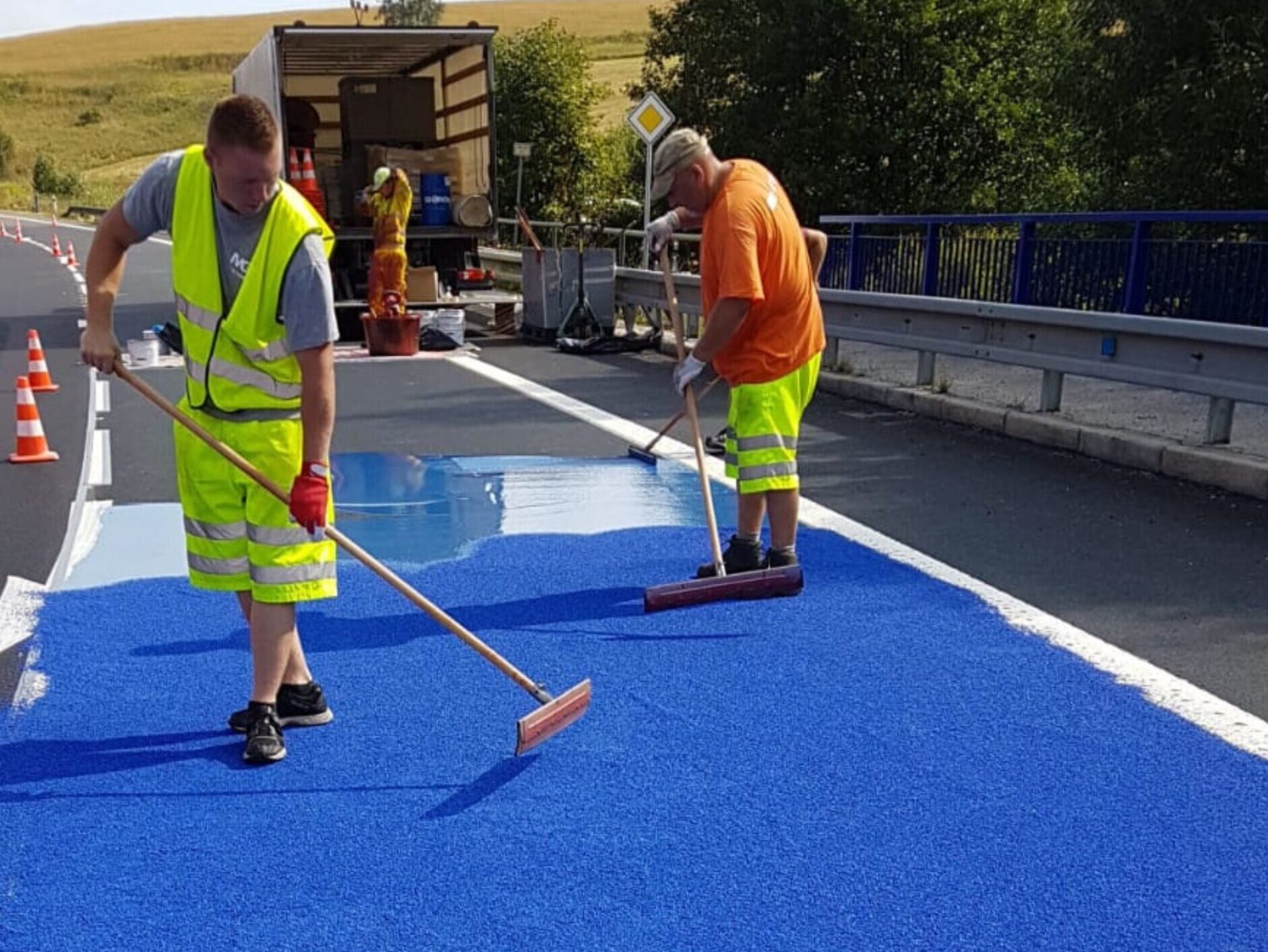Contractors applying blue Prismo Tyregrip high friction surfacing product with squeegees and spreading aggregate in Czechia